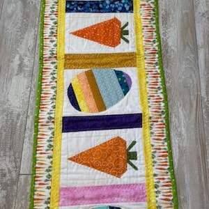 May include: A rectangular Easter-themed table runner. The runner features alternating carrot and egg designs in various colors, including orange, pink, and blue. The edges are trimmed with a green and orange carrot pattern, and the overall design is quilted.