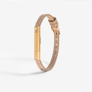 May include: A gold-toned metal bracelet with a beige leather band. The bracelet has a buckle closure and a small, round button on the front.