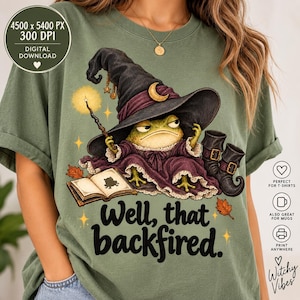 May include: An olive green t-shirt featuring a graphic of a green frog wearing a black witch hat and purple robe, holding a glowing wand over an open book. The text "Well, that backfired." is printed below the frog, surrounded by stars and leaves.