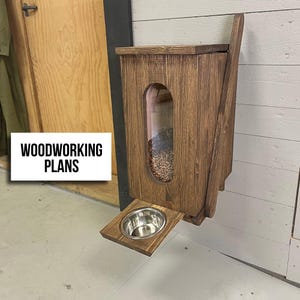 Peut inclure: Un distributeur de nourriture pour animaux en bois fixé au mur avec une finition brun foncé, doté d'une fenêtre verticale transparente montrant des croquettes à l'intérieur et d'une tablette rabattable avec un bol en acier inoxydable.