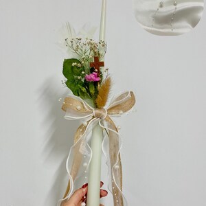Könnte beinhalten: Eine dekorative Kerze mit Blumenarrangement, Holzkreuz und großer Schleife. Die Kerze wird von einer Hand mit rotem Nagellack gehalten. Eine kleinere Haarschleife befindet sich in der oberen rechten Ecke. Die Kerze ist ca. 25 cm lang.