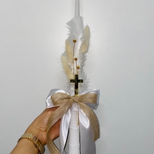 Puede incluir: Una vela blanca decorada con plumas blancas, flores amarillas secas y una cruz dorada. La vela está envuelta con cinta de raso blanco y un lazo de arpillera. Un artículo decorativo para ceremonias religiosas.