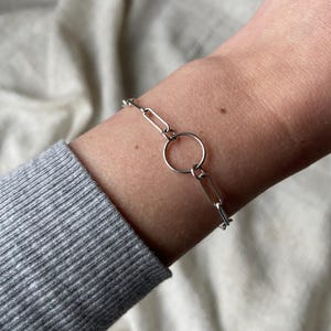 May include: A silver chain bracelet with a large circle link. The bracelet is on a person's wrist.