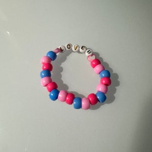 May include: A pink and blue beaded bracelet with the word "LOVE" spelled out in white beads with gold lettering. A pink heart bead with a white heart design is included in the bracelet.