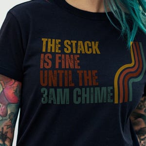 May include: Black t-shirt with a retro-style graphic that reads "The stack is fine until the 3AM chime".