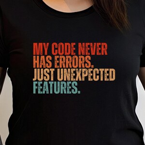 May include: A black t-shirt with a retro-style graphic that reads "My code never has errors. Just unexpected features."
