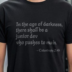 Op de afbeelding: Een zwart T-shirt met een witte grafische print die "In the age of darkness, there shall be a junior dev who pushes to main. -Catastrophe 2:404" zegt.