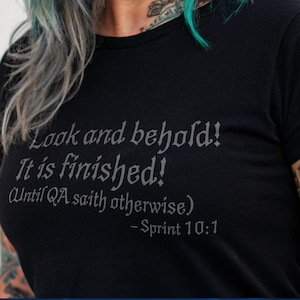 Può includere: Una maglietta nera con la scritta "Look and behold! It is finished! (Until QA saith otherwise) - Sprint 10:1". La persona che indossa la maglietta ha capelli colorati e tatuaggi. L'immagine include anche il testo "Unique Geek".