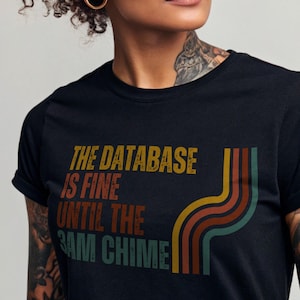 Può includere: Maglietta nera con una grafica in stile retrò che recita "The database is fine until the 3am chime".