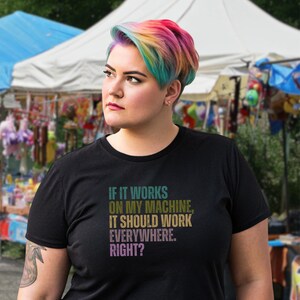Puede incluir: Camiseta negra con el texto "IF IT WORKS ON MY MACHINE, IT SHOULD WORK EVERYWHERE. RIGHT?" en verde, dorado y morado. La persona de la imagen tiene el pelo de color arcoíris.