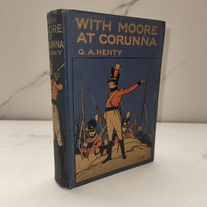 Può includere: Un libro vintage con copertina rigida intitolato "With Moore at Corunna" di G.A. Henty. La copertina è blu navy con scritte dorate e un'illustrazione colorata di soldati con cappotti rossi. Il libro è in buone condizioni.