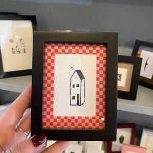 May include: A black framed print with a red and white checkered border. The print features a simple black and white illustration of a house with a bird flying above it.