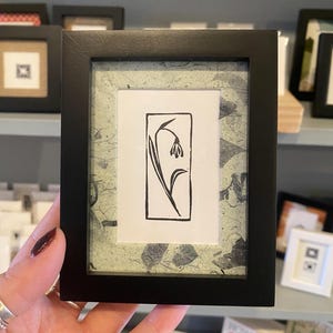 May include: A black and white line drawing of a flower in a black frame with a green paper mat. The flower is a single bloom with a stem and leaves.