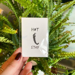 May include: A small white card with the words "HOT STUFF" and a black chili pepper illustration. The card is in a clear plastic sleeve, held against a backdrop of green foliage.