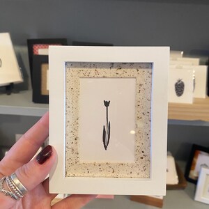 May include: A small white framed art print featuring a minimalist black tulip illustration. The frame has a speckled, textured border. The artwork is held in a hand with multiple silver rings. Other framed art pieces are visible in the background.