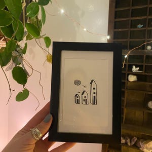 May include: A black framed print of a simple line drawing of three houses with a bird flying above them. The houses are different sizes and have a simple design.
