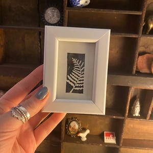 May include: A white frame with a black and white fern print inside. The frame is being held by a hand with silver rings.