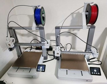 May include: Two white 3D printers with brown build plates and touch screens. One printer has a spool of blue filament and the other has a spool of red filament.