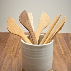 May include: A white ceramic utensil holder with a brown rim, filled with wooden cooking spoons.