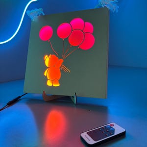 Könnte beinhalten: Ein quadratisches, beleuchtetes Kunstwerk mit einem Cartoon-Bären, der rosa Ballons hält. Der Bär und die Ballons leuchten orange und rosa. Eine Fernbedienung liegt vor dem Kunstwerk. Das Kunstwerk wird von einem Holzständer gehalten.