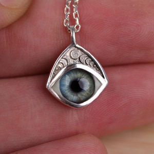 Puede incluir: Un collar con colgante de plata con un diseño detallado de ojo. El ojo tiene un iris azul y verde con una pupila negra, engastado en un marco de plata decorativo. La cadena es de plata.