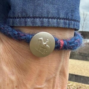 May include: A blue and red braided bracelet with a silver button featuring a raised image of a person riding a horse.