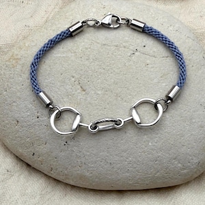 May include: A blue and silver bracelet with a horse bit design. The bracelet features a woven blue cord and silver-toned metal hardware, including a clasp and decorative elements. The bracelet is displayed on a light-colored stone.