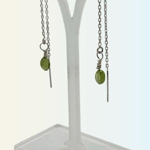 Pode incluir: Um par de brincos de corrente prateada com contas de pedras preciosas verdes penduradas na parte inferior. Os brincos são exibidos em um suporte de plástico transparente.
