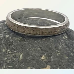 Könnte beinhalten: Ein silberner Ring mit einem strukturierten Band aus Sand. Der Ring liegt auf einem grauen Stein.