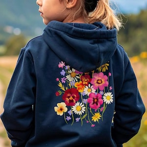 May include: A navy blue hooded sweatshirt with a colorful floral design on the back. The embroidered flowers include red, pink, yellow, and white blooms. The sweatshirt is worn by a child on a path in a field.