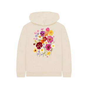 May include: Cream-colored hoodie with a floral design on the back. The floral arrangement features a variety of colorful flowers, including purple pansies, pink daisies, and yellow sunflowers. The hoodie has a hood and long sleeves.