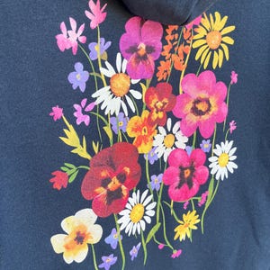 May include: Navy blue hoodie featuring a vibrant floral design on the back. The bouquet includes pink, red, yellow, and white flowers with green stems. The flowers are clustered together, creating a colorful and eye-catching design.