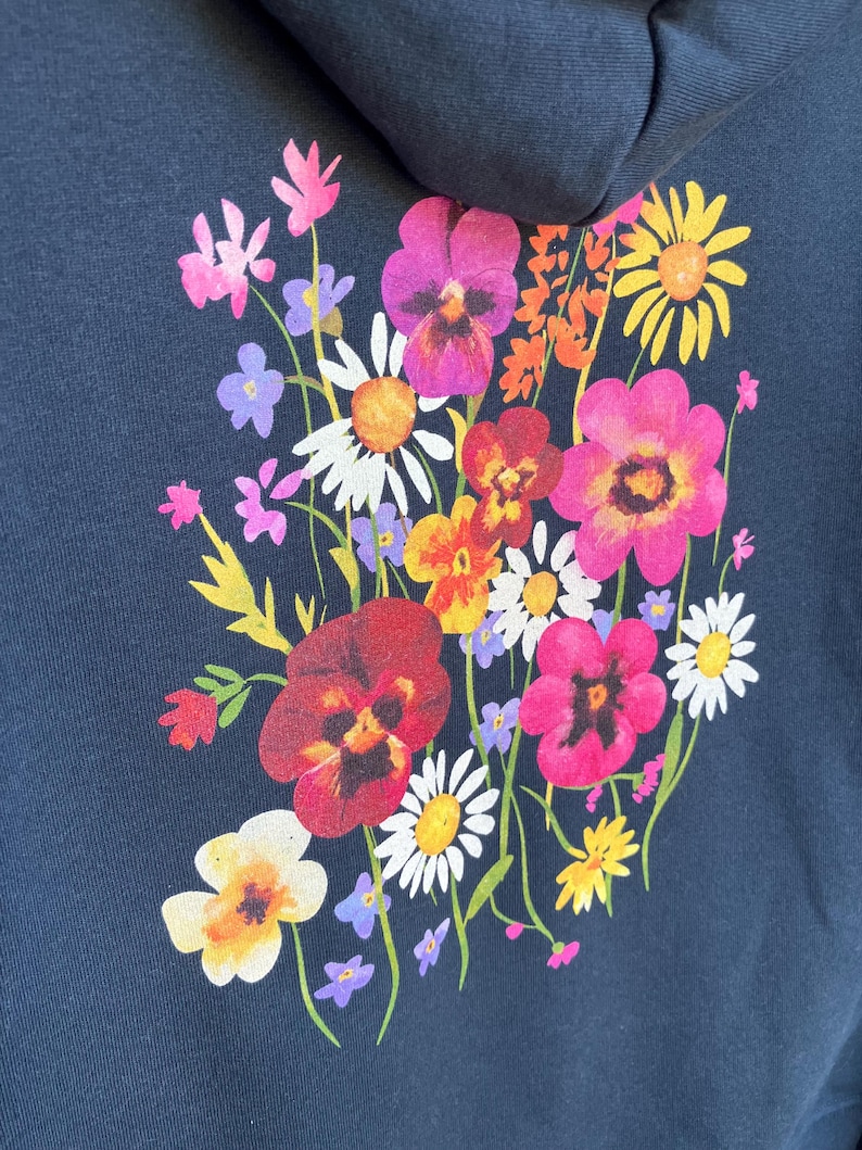 May include: Navy blue hoodie featuring a vibrant floral design on the back. The bouquet includes pink, red, yellow, and white flowers with green stems. The flowers are clustered together, creating a colorful and eye-catching design.