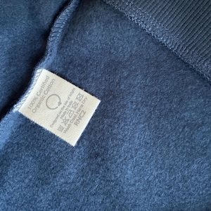 May include: Close-up of a navy blue fabric with a white tag. The tag reads "100% Certified Organic Cotton" and includes care instructions. The fabric has a soft, textured appearance, with a ribbed section visible.