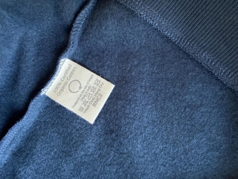 May include: Close-up of a navy blue fabric with a white tag. The tag reads "100% Certified Organic Cotton" and includes care instructions. The fabric has a soft, textured appearance, with a ribbed section visible.