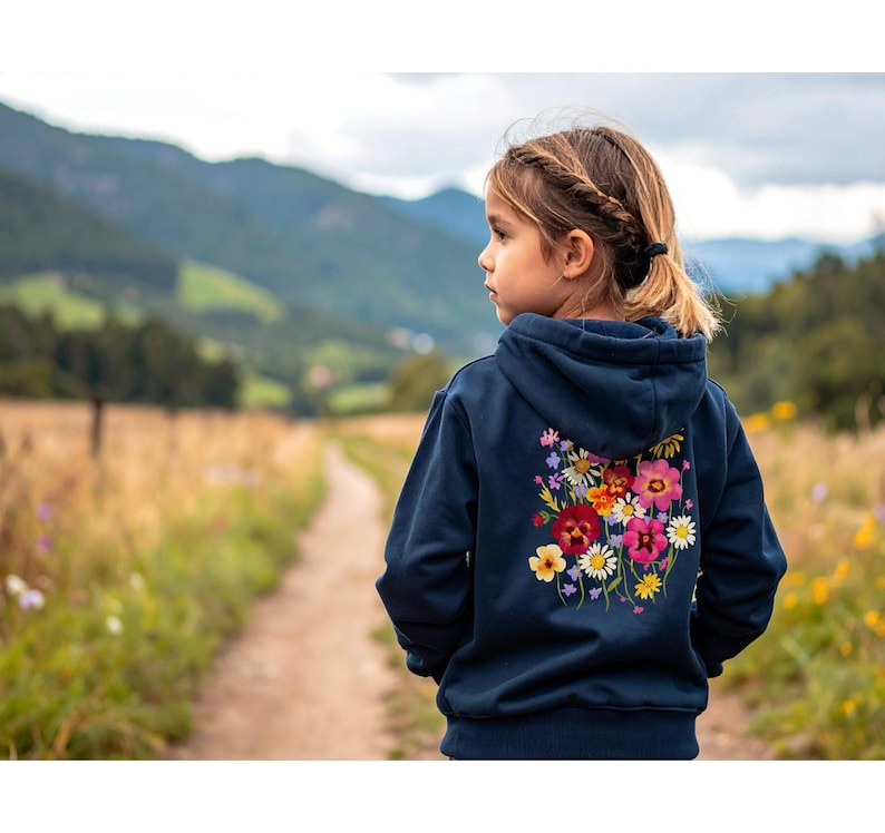 May include: A navy blue hooded sweatshirt with a colorful floral design on the back. The embroidered flowers include red, pink, yellow, and white blooms. The sweatshirt is worn by a child on a path in a field.
