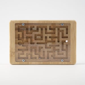 May include: A wooden maze puzzle with a clear plastic top. The maze is carved into the wood and contains a small metal ball. The wooden frame is rectangular and secured with four screws. The maze is a light brown color.