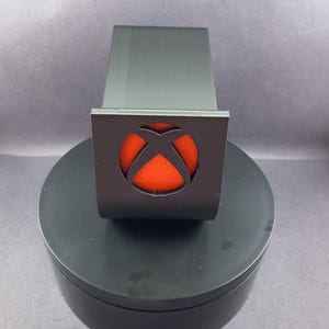 May include: A black plastic stand with a red Xbox logo cutout. The stand is designed to hold a gaming console.