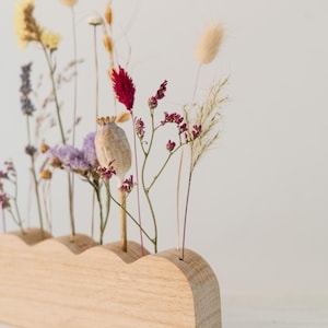 May include: A wooden vase with a wavy top holds a collection of dried flowers. The flowers include yellow, purple, and red blooms, along with fluffy beige grasses. The vase is made of light-colored wood.