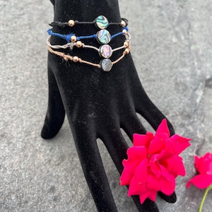May include: Four bracelets with abalone shell beads and gold-colored accents are displayed on a black hand-shaped display. The bracelets are black, blue, and rose-colored. A bright pink rose is placed near the base of the display.