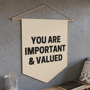 May include: A beige canvas banner with the text "YOU ARE IMPORTANT & VALUED" printed in black. The banner is hanging from a wooden dowel rod with twine.