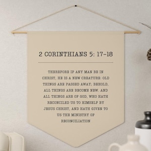 May include: Beige canvas wall hanging with a gold rod at the top. The canvas features black text that reads "2 Corinthians 5:17-18" and a bible verse about being a new creature in Christ.