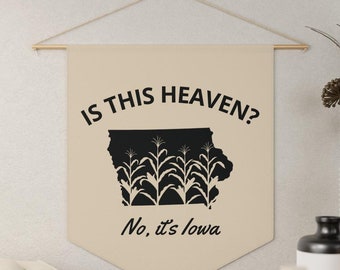 May include: A beige canvas wall tapestry with the text "Is this heaven?" and a black silhouette of the state of Iowa with corn stalks inside. The text "No, it's Iowa" is below the state outline.