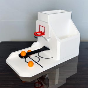 May include: White desktop organizer with a basketball hoop and a black ramp for rolling orange balls into the hoop. The organizer has a compartment for pens and pencils.