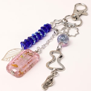 May include: A decorative keychain with a silver clasp and various charms. It features a pink rectangular bead, a strand of blue beads, a silver leaf, a silver hourglass shape, and a silver sandal charm. The background is white.