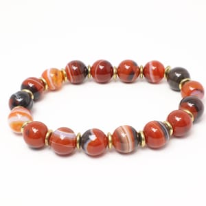 May include: A bracelet made of polished, round beads in red, orange, and brown hues. The beads are separated by small gold-coloured spacers. The bracelet is presented against a white backdrop.