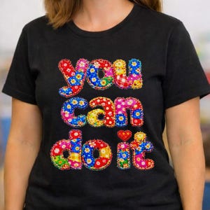 Puede incluir: Camiseta negra con las palabras "You can do it" en letras coloridas con estampado floral. La camiseta tiene cuello redondo y mangas cortas. El diseño presenta colores rojo, azul, amarillo y rosa.