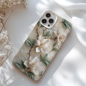 May include: A white phone case with a floral design featuring white flowers and green leaves. The case has a camera lens cutout with a silver ring around it.