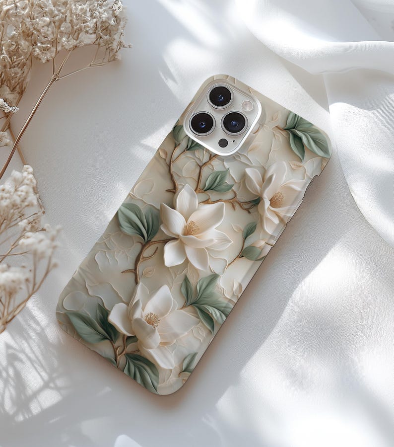 May include: A white phone case with a floral design featuring white flowers and green leaves. The case has a camera lens cutout with a silver ring around it.
