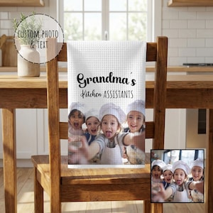 May include: White waffle-weave kitchen towel with the text "Grandma's Kitchen Assistants" in black script. The towel features a photo of children wearing chef hats and aprons. The towel is hanging on a wooden chair.